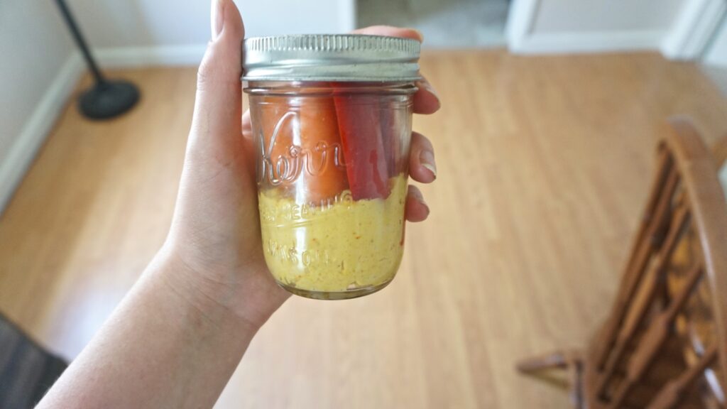 a hand holding a glass jar with hummus and bell peppers