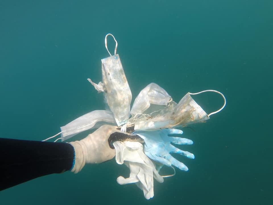 a hand in the ocean holding disposable face masks and plastic gloves