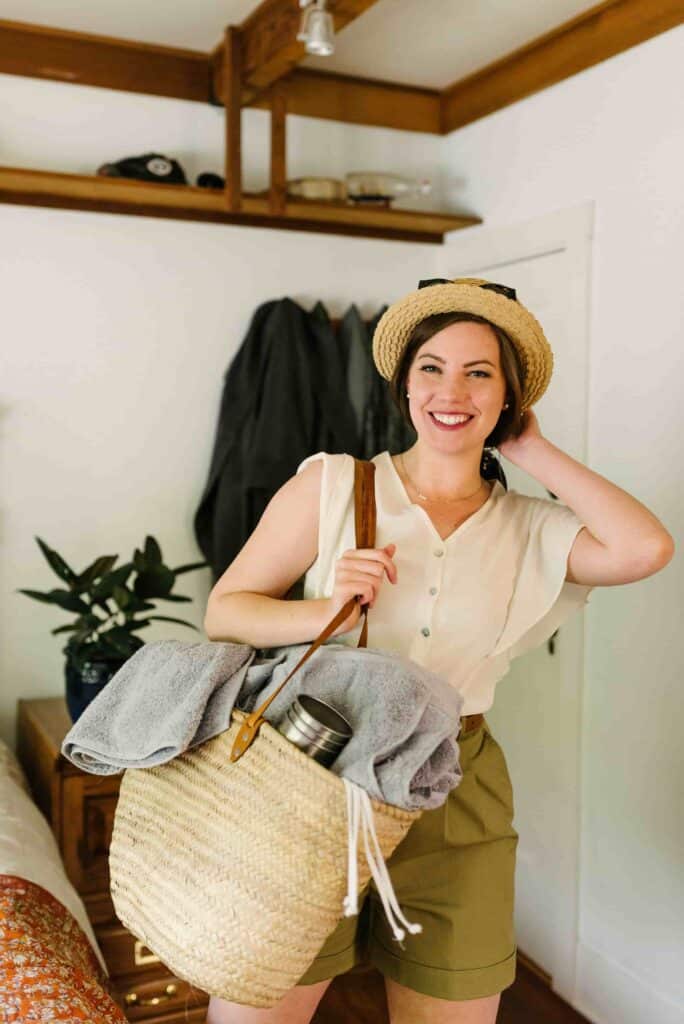 Smiling woman with her beach necessities - sun hat, sun glasses, beach bag, stainless steel cup, and towel.