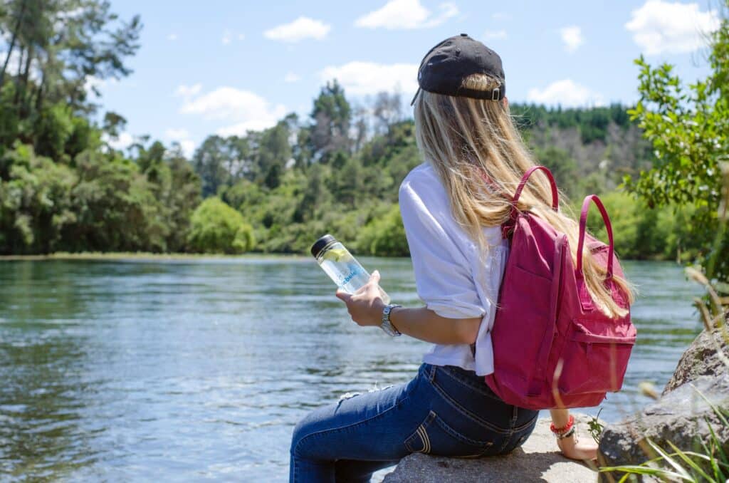 Woman sitting on a rock wearing a backpack and hat, holding a glass water bottle.