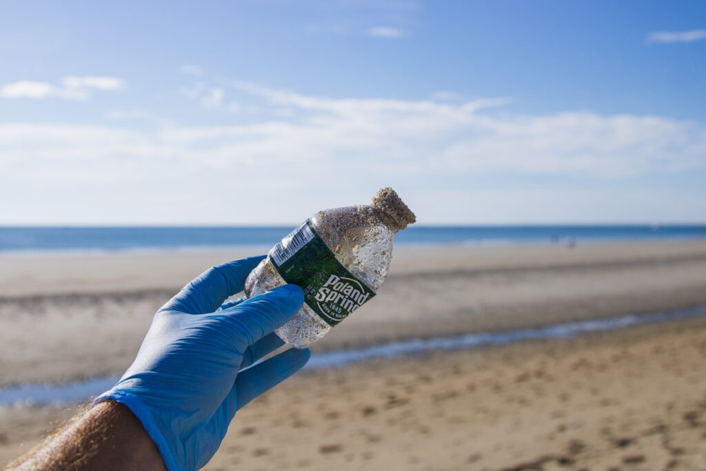 A hand holding a single use plastic water bottle discarded at the beach showing the importance of recycled water bottles. 