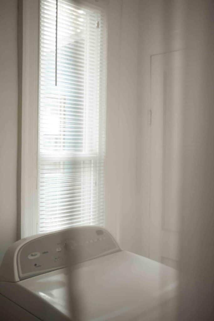 A white washing machine in a white room with a window. 