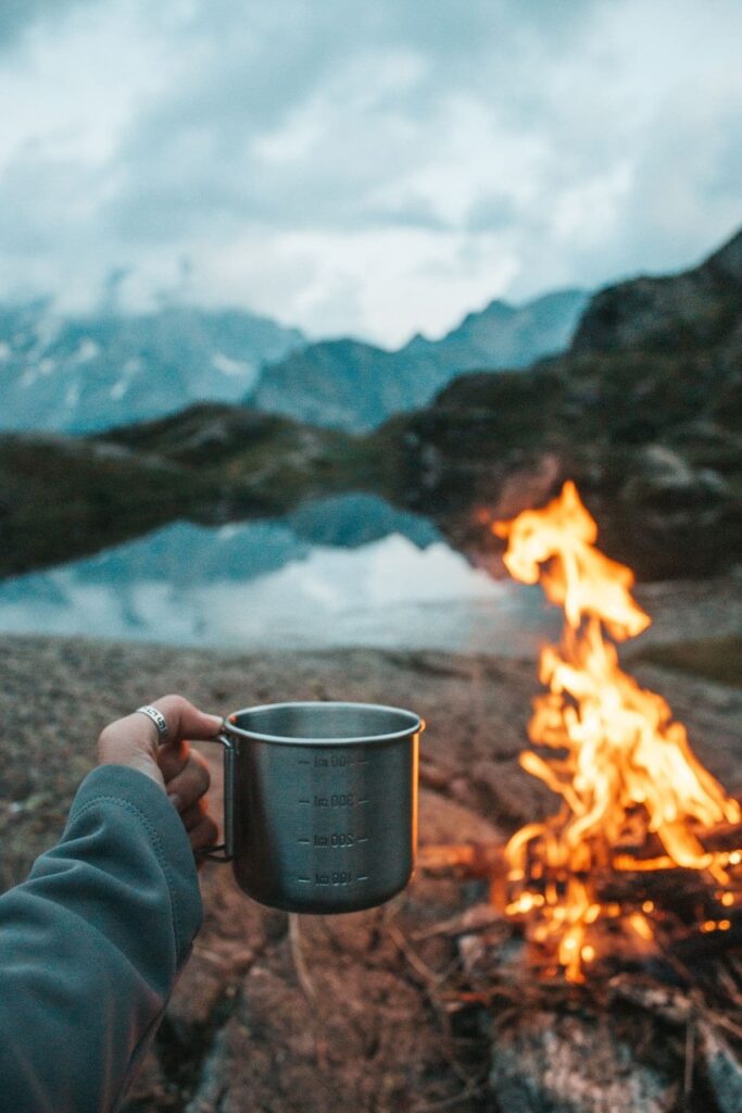 A woman holding a tin cup in front of a campfire to illustrate websites like Amazon for outdoor gear.